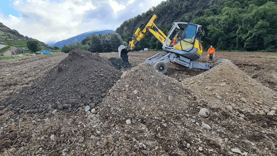 Une petite pelle mécanique jaune creusant un champs en bord de route dans une vallée.