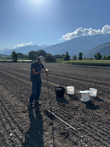 Un homme dans un champs labouré avec une sonde en mains plantée dans le sol.