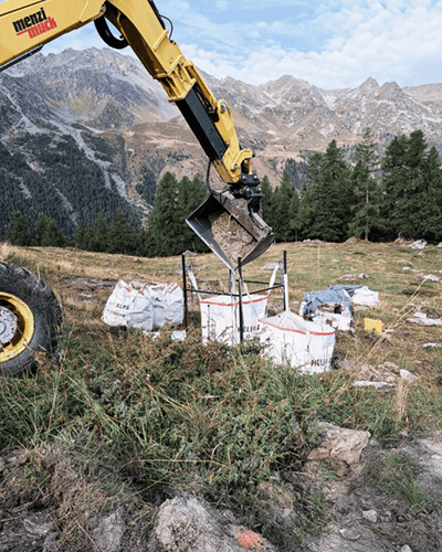 Une pelle mécanique jaune assainissant un stand de tir en montagne et remplissant des sacs de plastic blancs de terre.