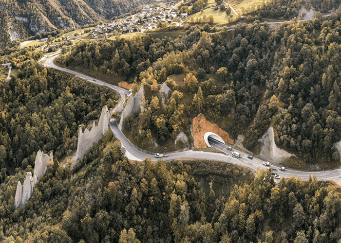 Vue aérienne d'un petit tunnel alpin, entouré de sapin ainsi que de l'ancienne route qui contournait la montagne.