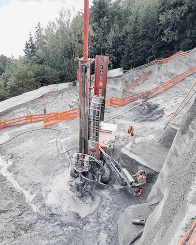 Une grande foreuse entourée de résidus de roche avec des sapins en fond.
