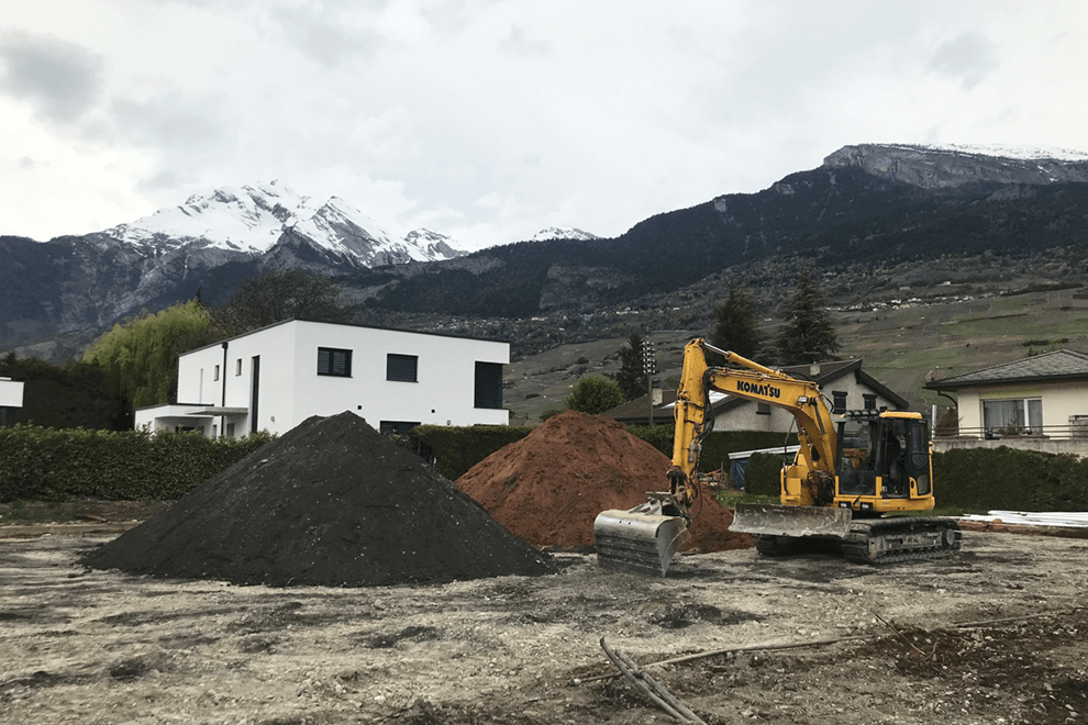 Une pelle mécanique jaune sur la droite et deux tas de mâchefer sur la gauche, avec des villas et les Alpes en fond.