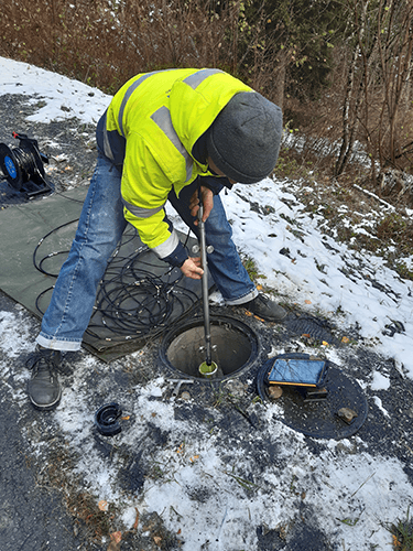 Un ingénieur du Bureau BEG portant une veste réfléchissante jaune et soulevant un instrument de mesure ressemblant à un Karcher au dessus d'une bouche d'égout ouverte.