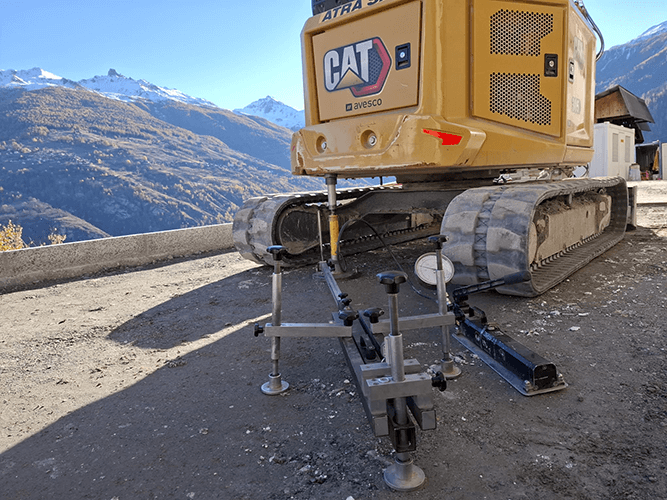 Une pelle mécanique jaune vue de près de derrière avec les Alpes automnales en fond. La pelle mécanique a un instrument de mesure installé à l'arrière.